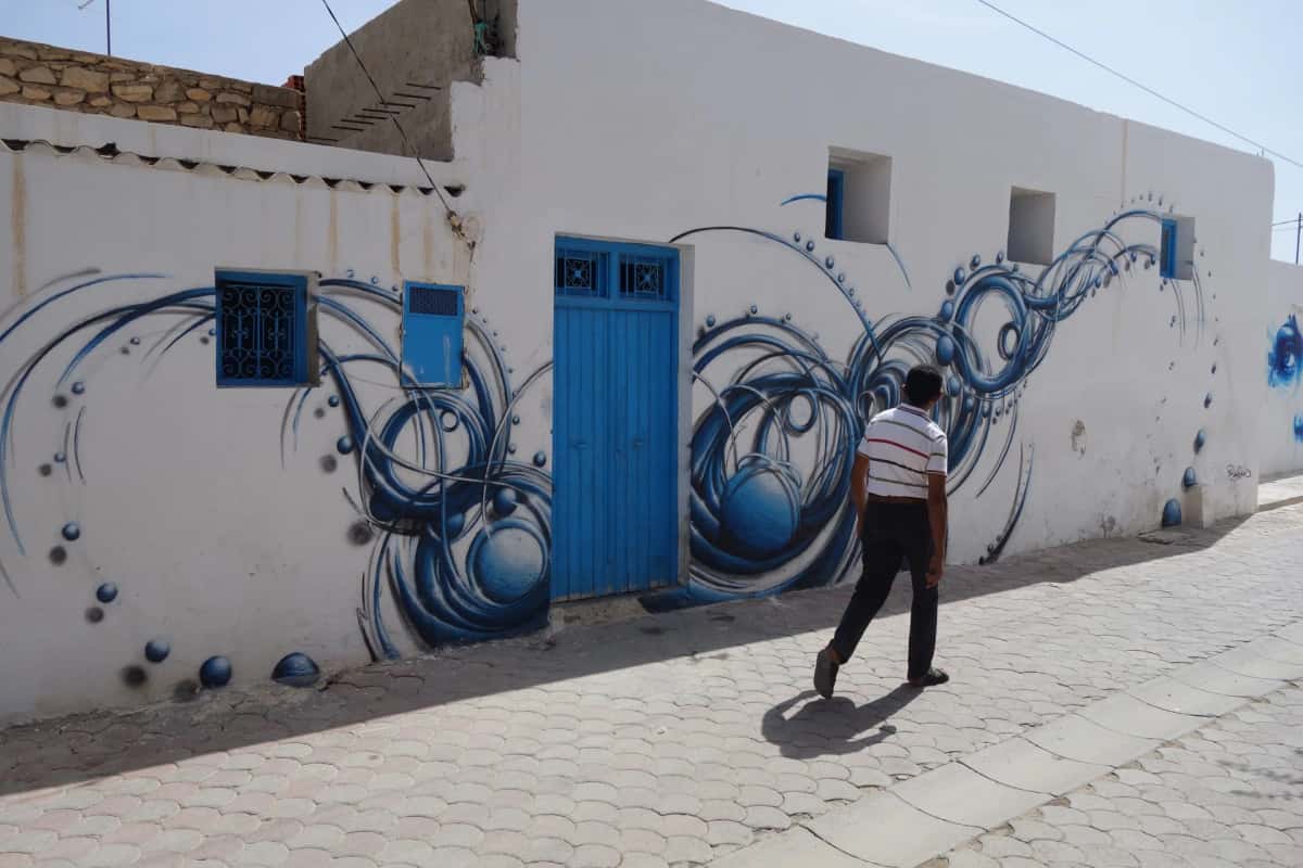 Passant devant une fresque bleue aux volutes sur une façade blanche à Erriadh, Djerbahood, Djerba