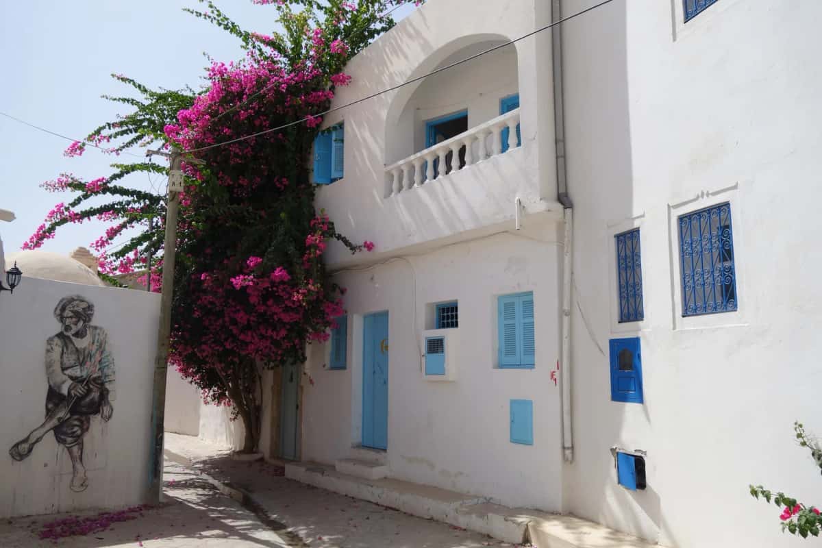 Ruelle blanche aux volets bleus et bougainvillées avec un portrait peint, à Erriadh, Djerba