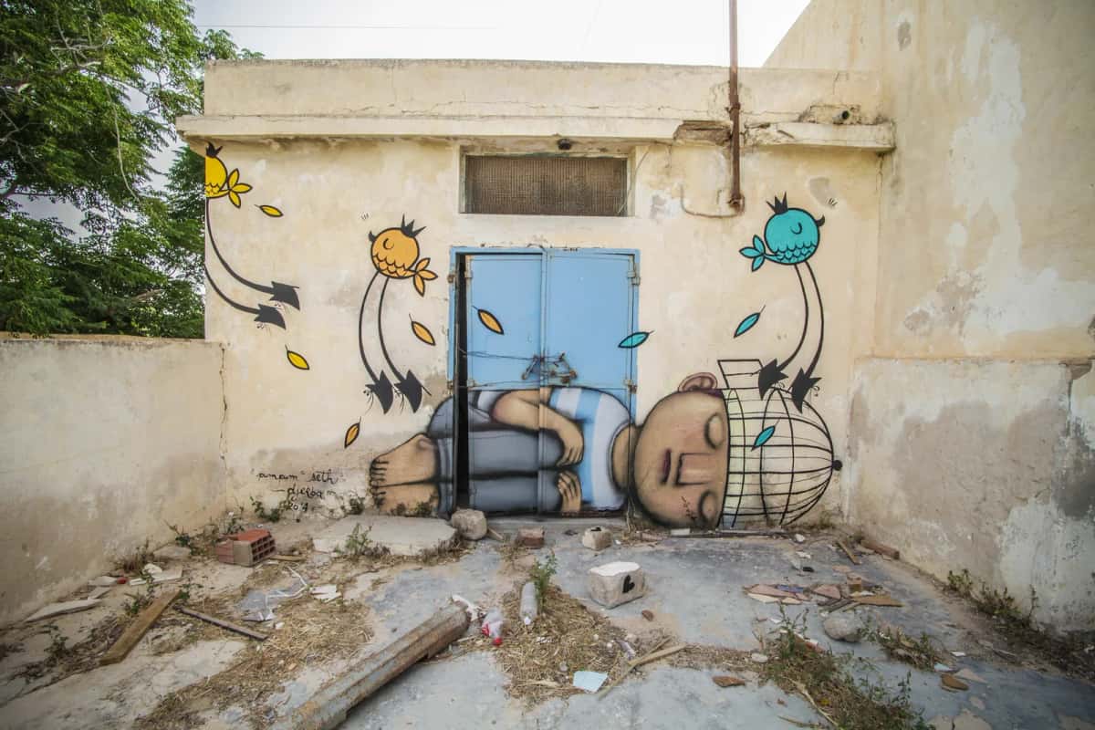 Enfant endormi peint par Seth avec Pum Pum devant une porte bleue à Erriadh, Djerbahood, Djerba