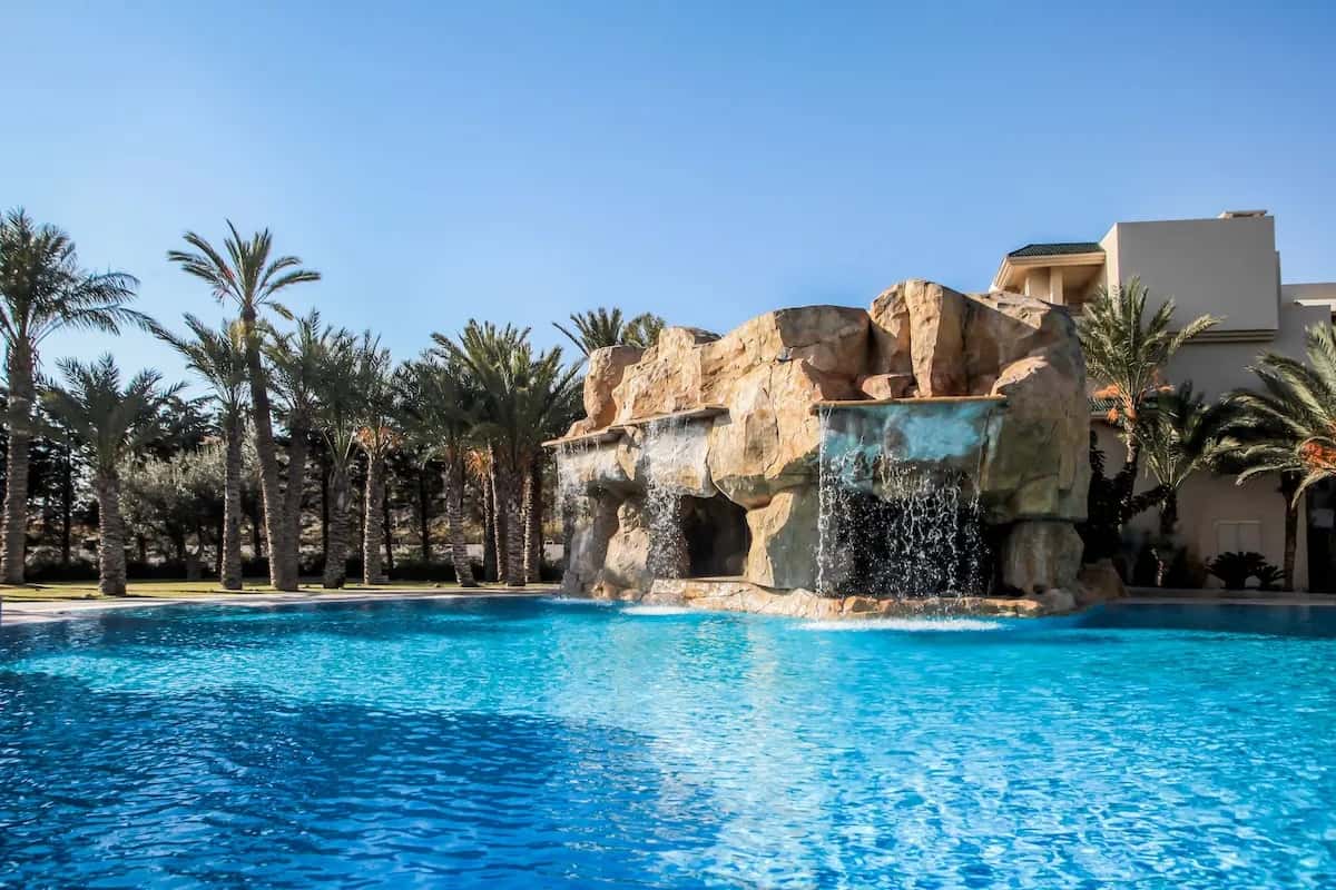Piscine avec cascade de rochers et palmiers sous un ciel bleu à The Russelior Hotel & Spa, Yasmine Hammamet